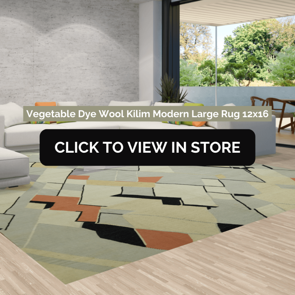 A modern open-concept living room showcasing a large, abstract-patterned Kilim rug in muted green, black, and orange tones. The rug lies beneath a white sectional sofa, with a floor-to-ceiling window offering a view of lush greenery outside.