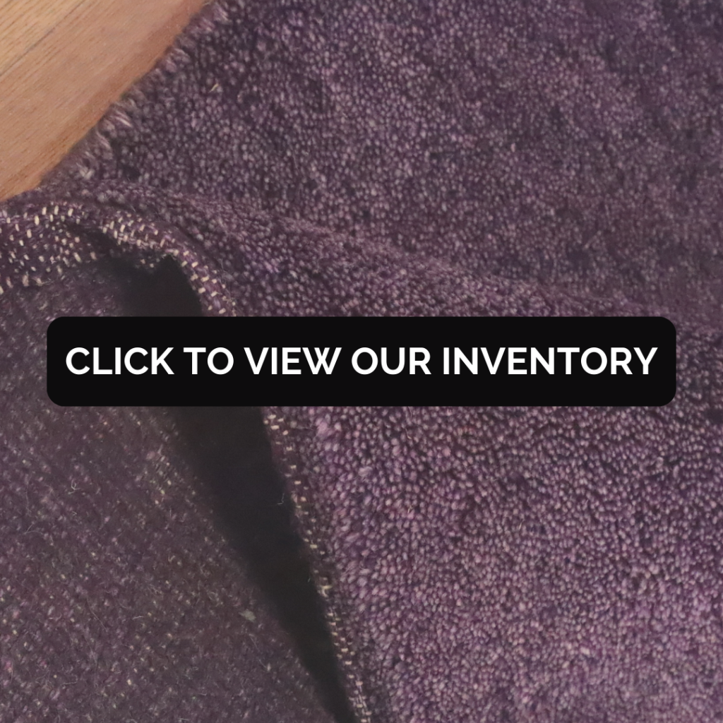 A close-up of a deep purple rug with a rich, textured weave, showing a folded corner. Text overlay reads “Click to view our inventory.”