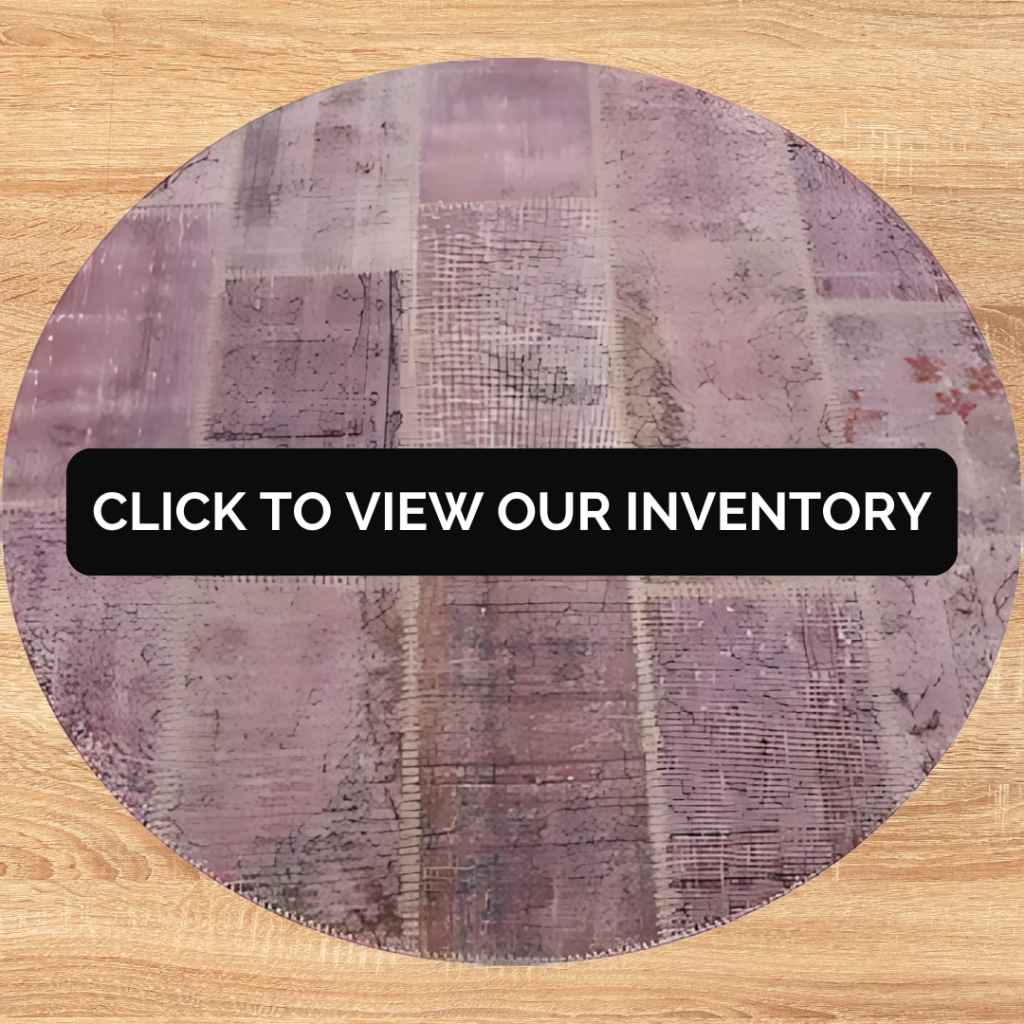 A round lavender-toned rug with abstract patchwork patterns displayed on a light wood floor. Text overlay reads “Click to view our inventory.”