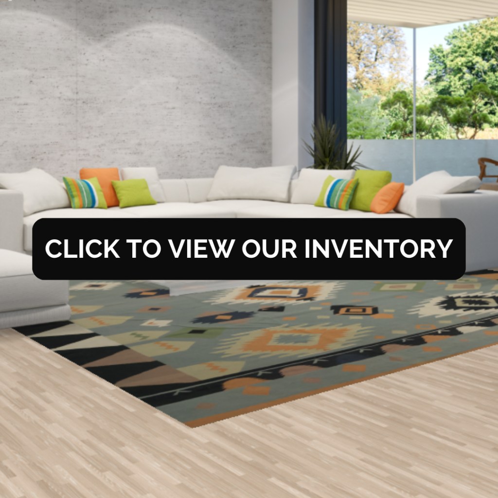 A bright modern living room with a white sectional sofa decorated with colorful throw pillows, sitting on a geometric patterned rug. Large windows open to a lush green outdoor view. Text overlay reads “Click to view our inventory.”