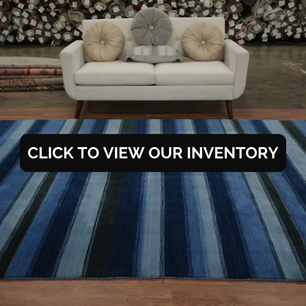 A striped blue rug placed in front of a cream-colored loveseat with round accent pillows, set against a backdrop of stacked rolled rugs. Text overlay reads “Click to view our inventory.”