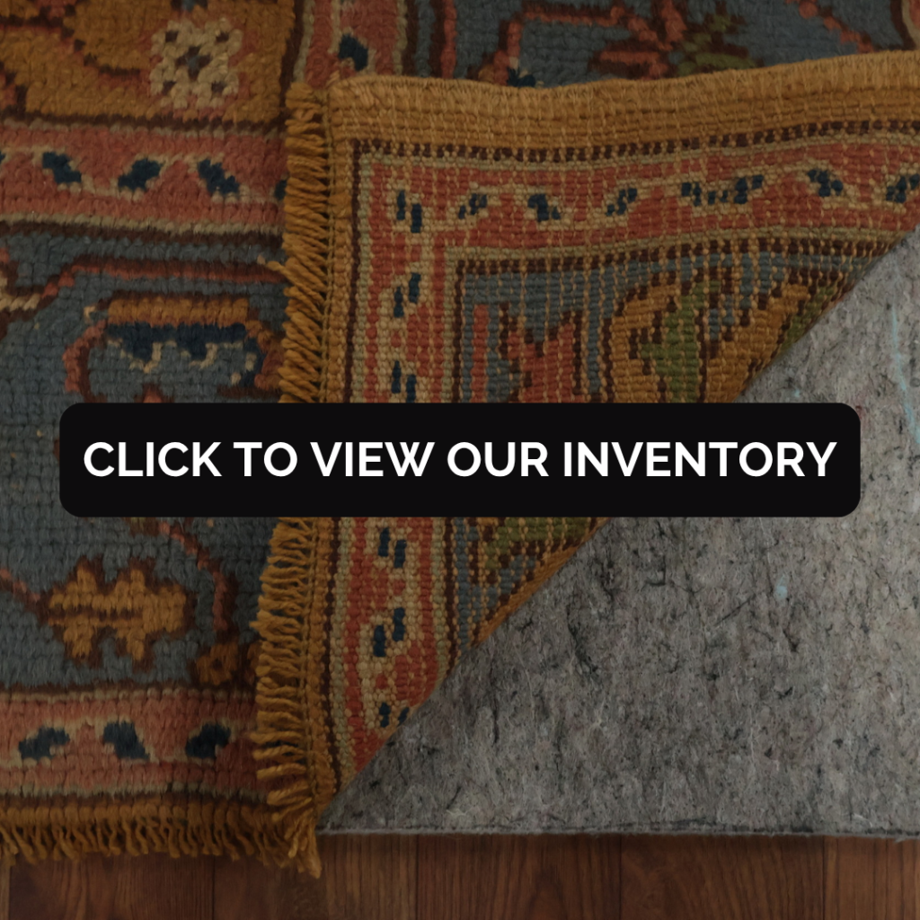 A close-up of a traditional woven rug in muted blue and rust tones with a golden border. One corner is folded over to reveal the backing and detailed fringe. Text overlay reads “Click to view our inventory.”