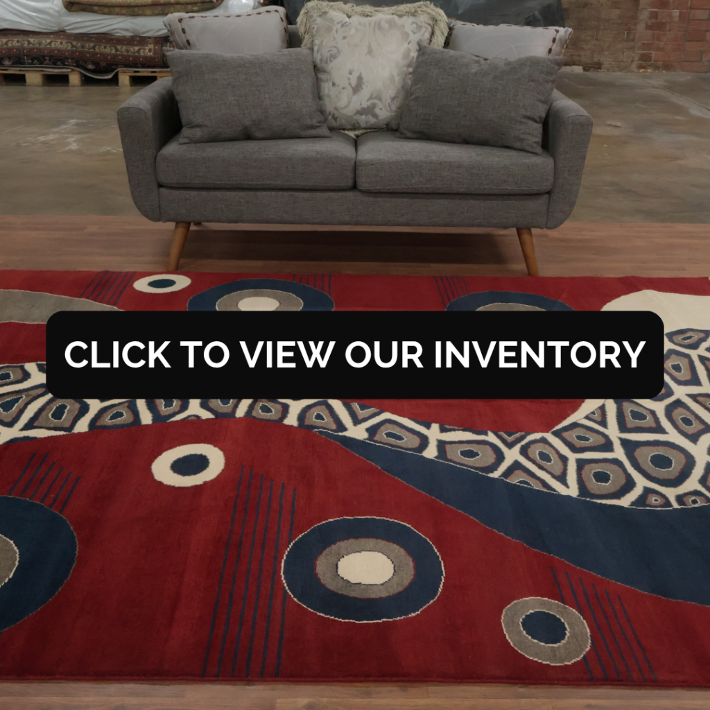 A bold rug in deep red with abstract circular patterns in navy, cream, and gray, styled in front of a gray loveseat. Text overlay reads “Click to view our inventory.”