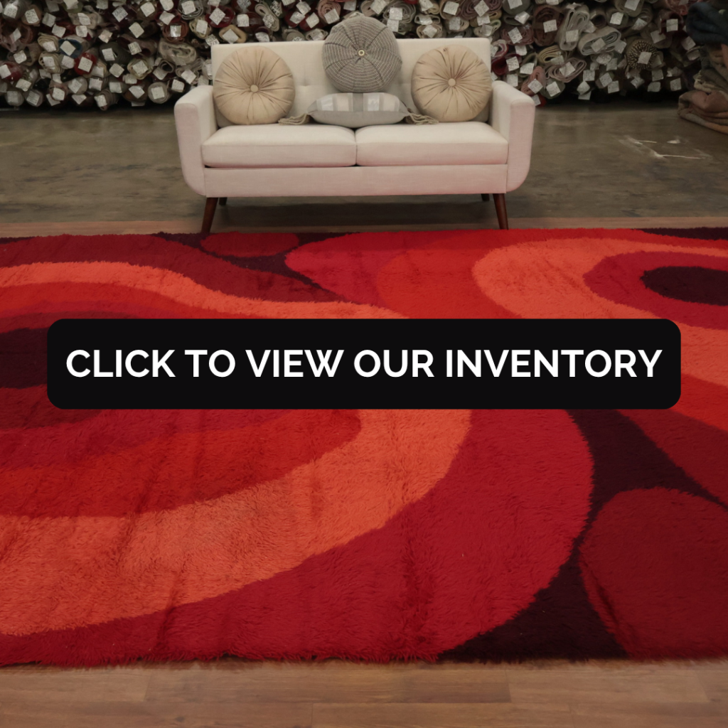 A modern beige couch with round pillows sits behind a bold red and orange shag rug featuring wavy, abstract patterns. Rolls of carpet line the background, and a black overlay in the center reads, “Click to view our inventory.”