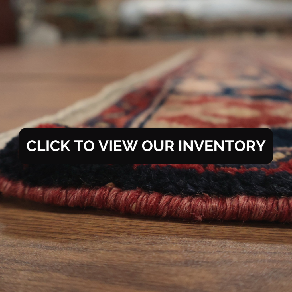 A close-up, side view of a handwoven rug with red, navy, and beige details lying on a wooden floor. A black banner across the center reads, “Click to view our inventory.”