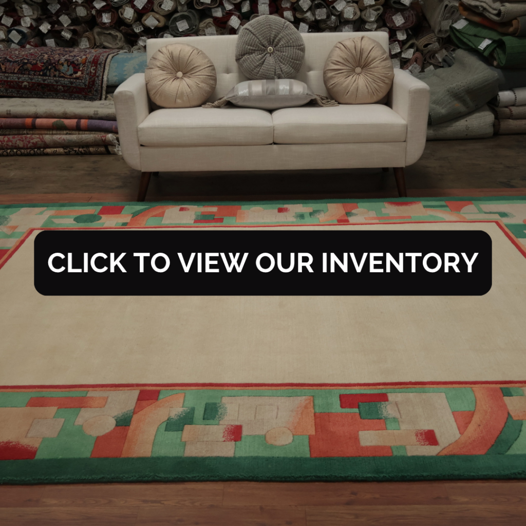 A cream sofa with round pillows sits atop a large beige rug bordered by abstract geometric patterns in green, red, and peach. Rolled rugs fill the background of the warehouse setting.