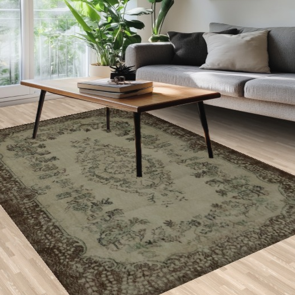 A close-up of a cozy living area with a wooden coffee table, gray sofa, and an antique-style green rug with ornate floral detailing and a dark border, illuminated by natural sunlight.