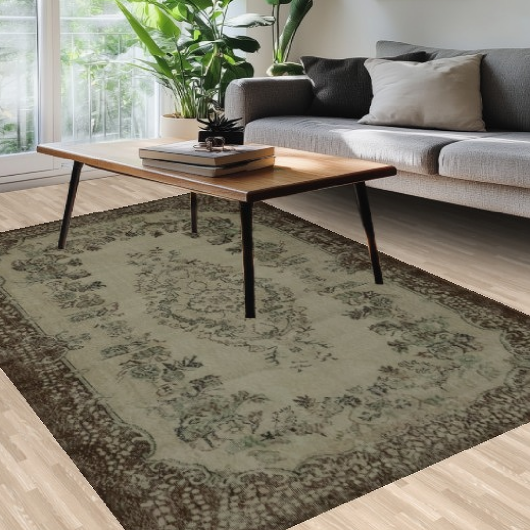 A close-up of a cozy living area with a wooden coffee table, gray sofa, and an antique-style green rug with ornate floral detailing and a dark border, illuminated by natural sunlight.
