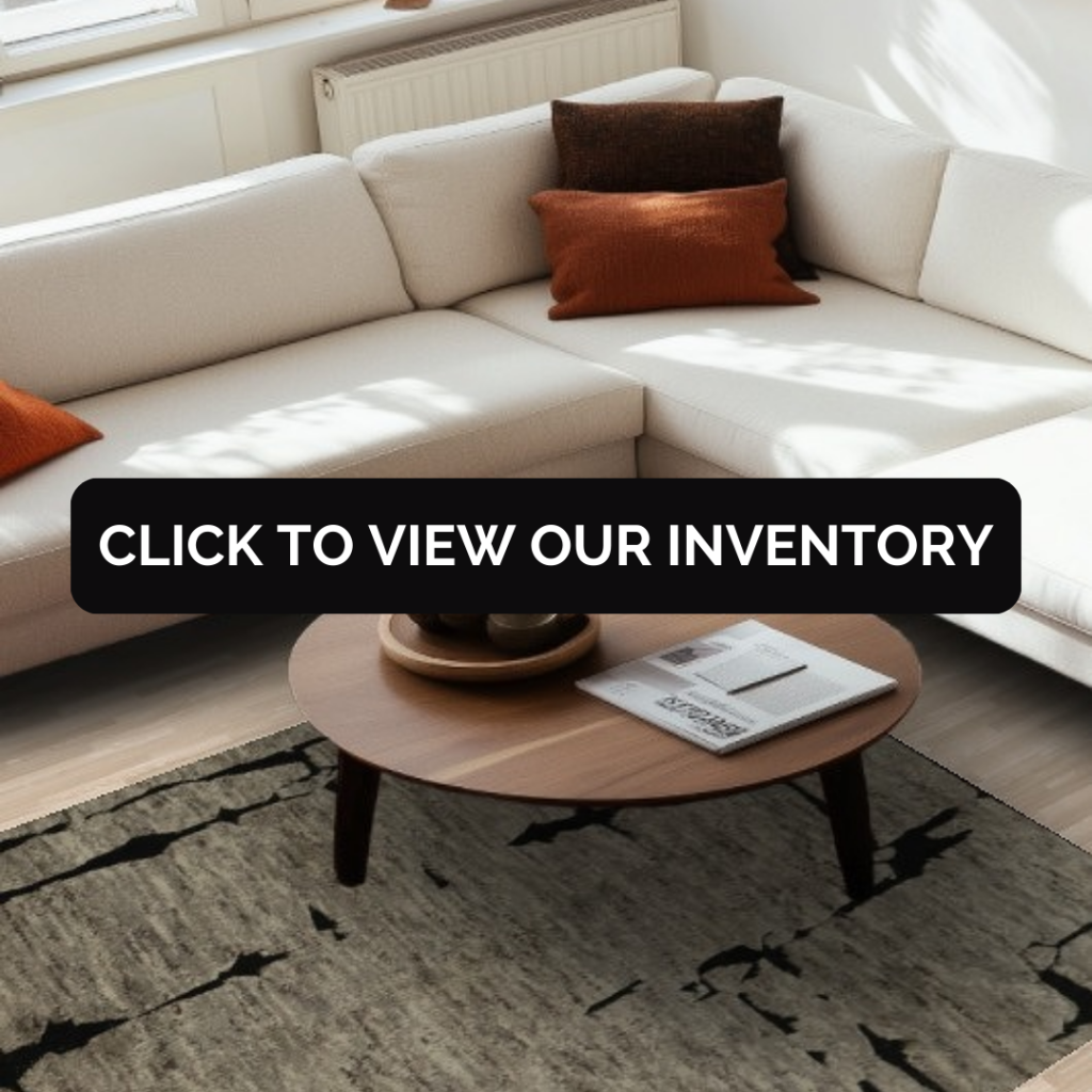 A bright living room features a cream-colored sectional sofa accented with rust and brown throw pillows. A round wooden coffee table sits on top of a textured rug with an abstract black-and-beige pattern. A black button with the text “CLICK TO VIEW OUR INVENTORY” is centered over the image.