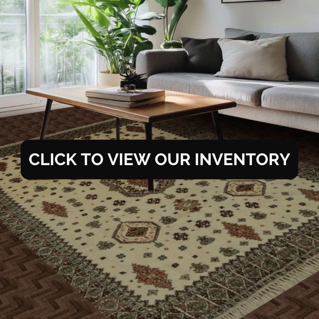 A modern living room shows a gray upholstered sofa with neutral throw pillows beside a wall of large windows and lush green plants. A light cream rug with intricate traditional motifs sits beneath a long wooden coffee table. A black “CLICK TO VIEW OUR INVENTORY” button overlays the center of the rug.