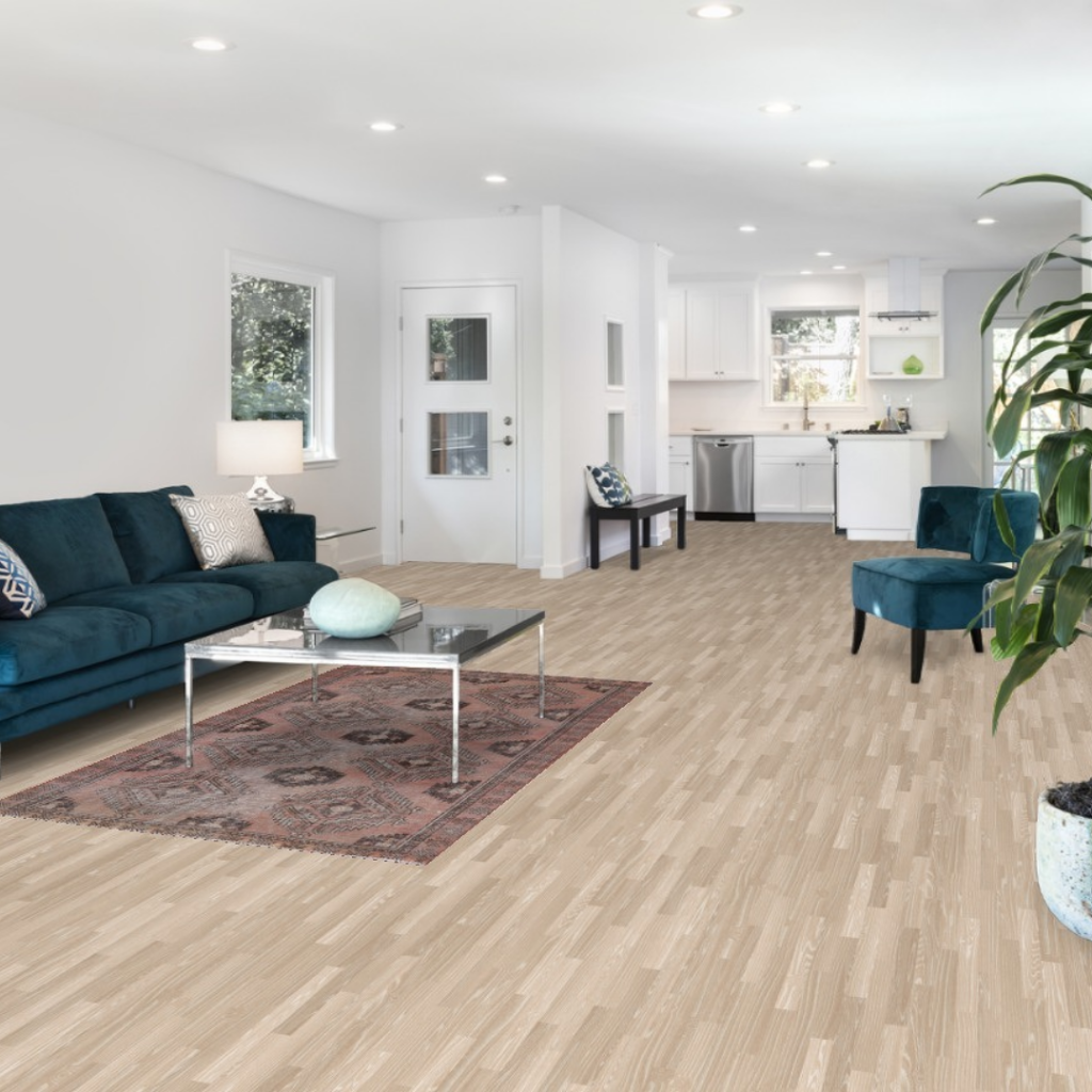 A spacious open living area features a teal velvet sofa and a glass coffee table positioned over a faded red patterned rug. The light wood floors and white kitchen create a clean, modern backdrop. Natural light brightens the entire space, giving it an inviting feel.