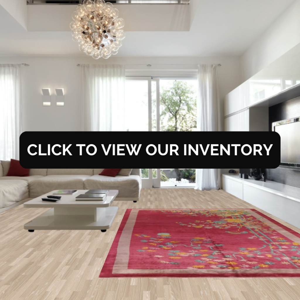 A contemporary living room with white furnishings and large windows showcases a striking pink rug with colorful floral details. A low white coffee table sits centered on the rug beneath a dramatic chandelier. A black “CLICK TO VIEW OUR INVENTORY” button is displayed across the middle.