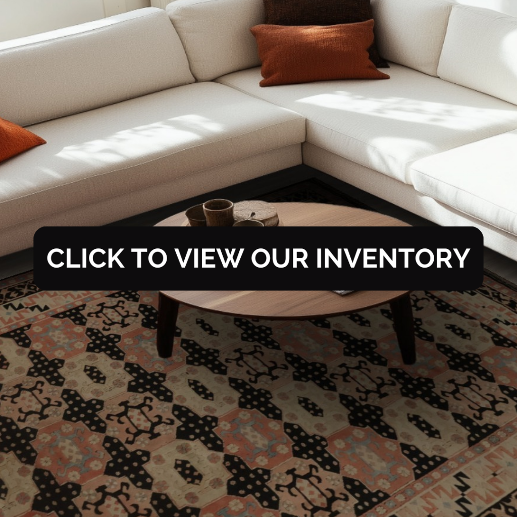 A minimalist living room features a cream-colored sofa with a black-and-white patterned pillow beside a small black accent table. A large Southwest-inspired rug in shades of coral, green, and black anchors the space. A black button with “CLICK TO VIEW OUR INVENTORY” is displayed across the rug.