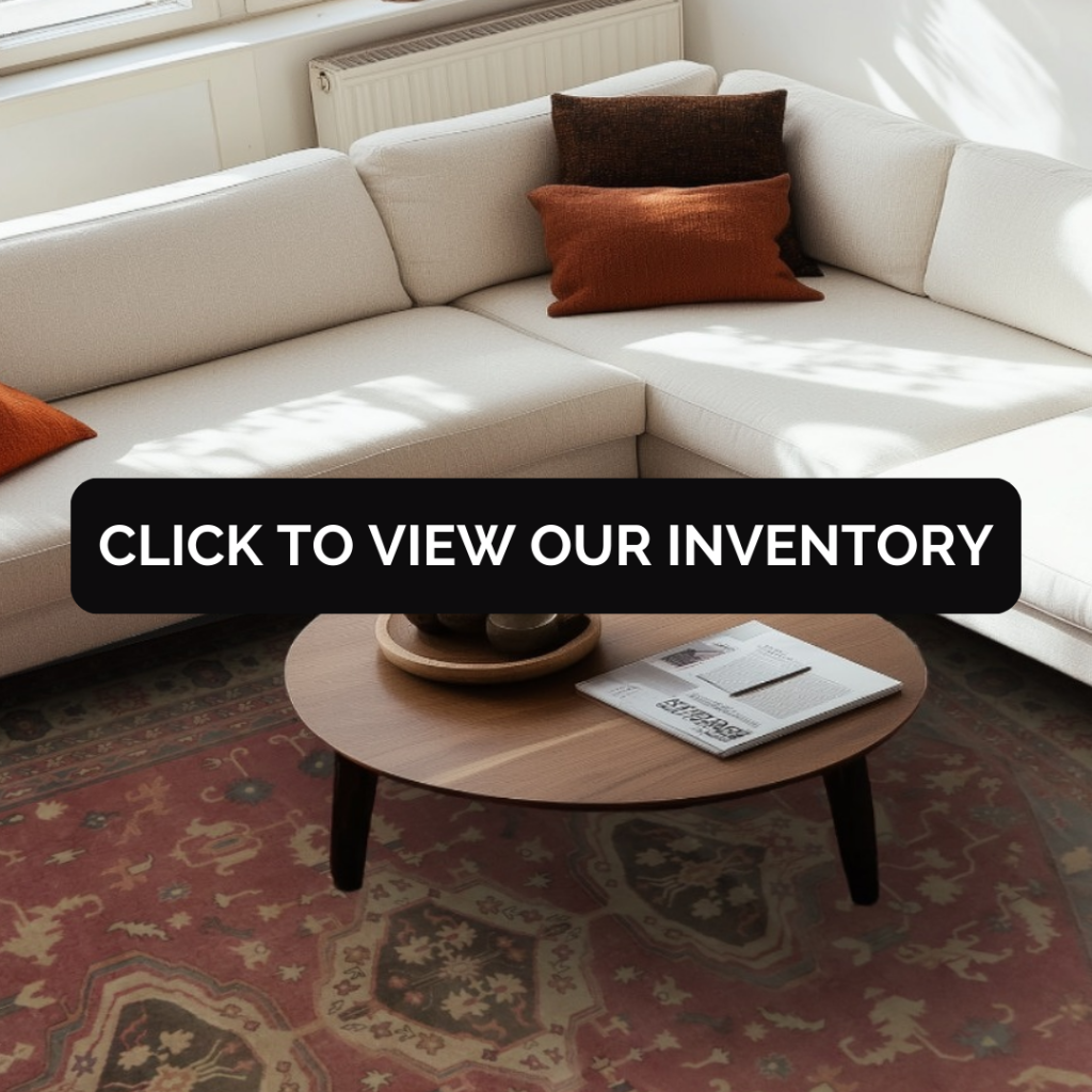 A cream-colored sectional sofa with warm-toned throw pillows sits in a sunlit living room. A muted rose rug with soft geometric patterns extends beneath a round wooden coffee table. A black overlay button reads “CLICK TO VIEW OUR INVENTORY.”