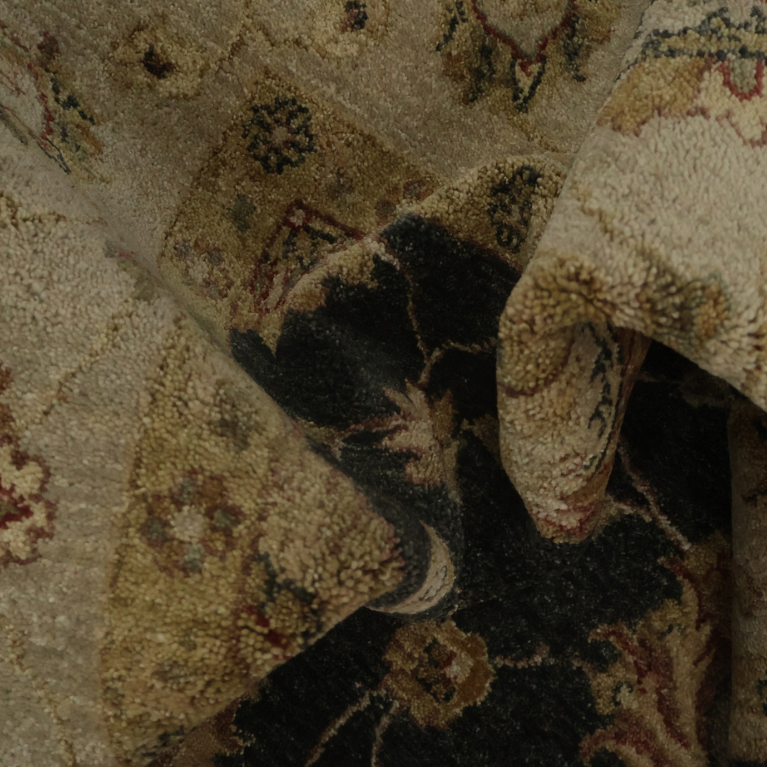 A tightly framed close-up shows a thick, folded rug with ornate traditional patterns in earthy tones. The pile appears dense and textured, highlighting the rug’s handwoven detail. Soft lighting emphasizes the contrast between lighter beige areas and dark brown sections.