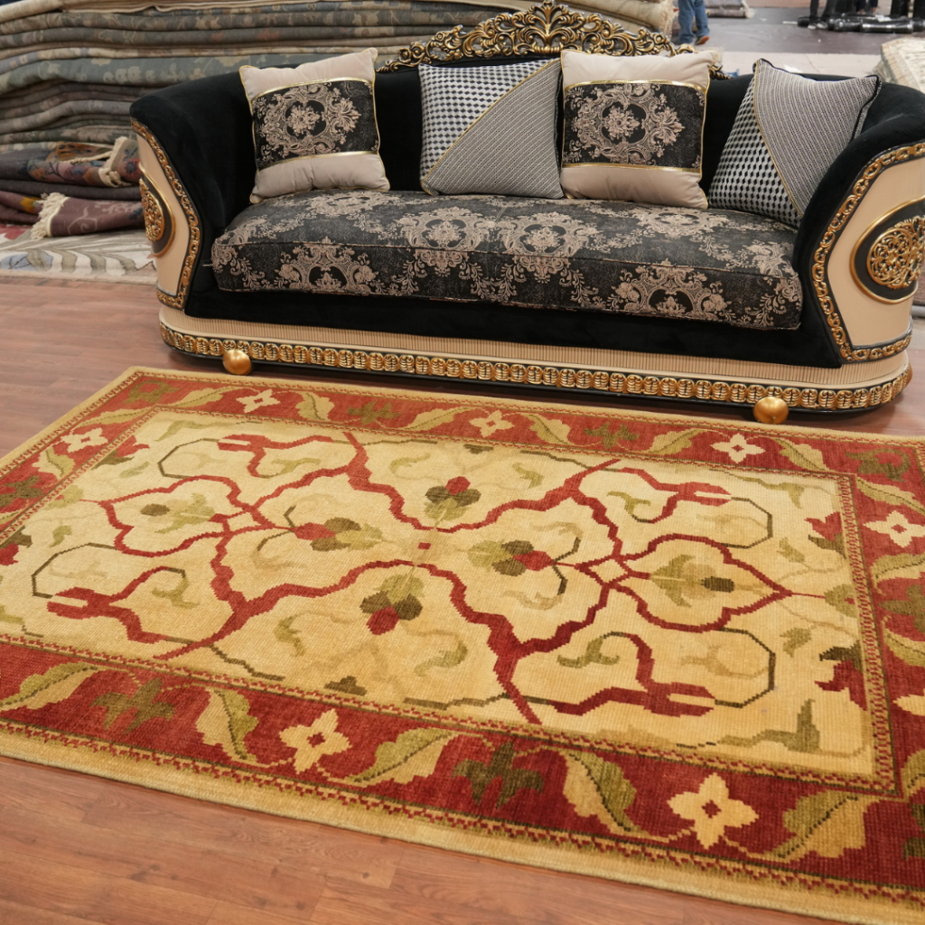 A lavish black-and-gold sofa with intricately patterned cushions sits on a hardwood floor in a rug showroom. A large red and yellow floral rug fills the foreground, complementing the sofa’s ornate detailing. Rolled rugs are stacked behind the display, suggesting a showroom environment.
