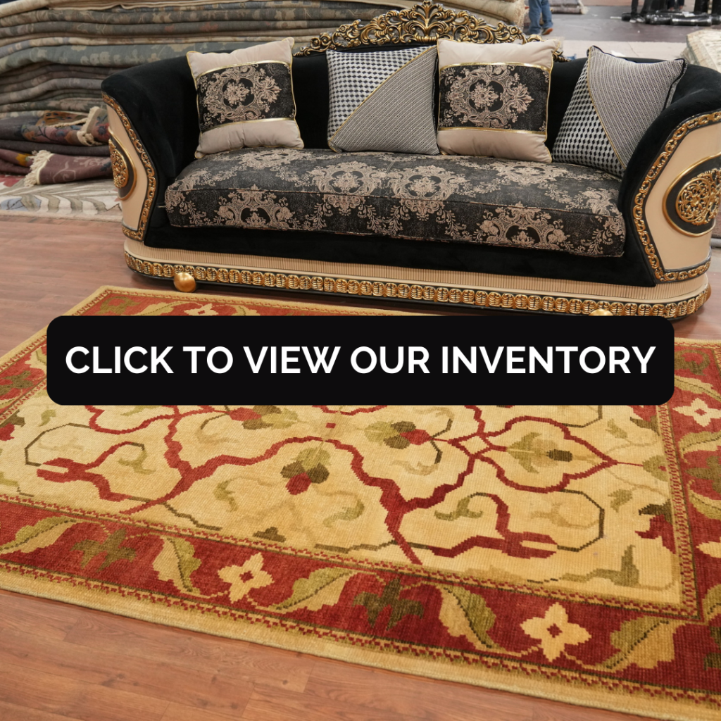 A luxurious black-and-gold upholstered sofa with decorative pillows is displayed in a showroom surrounded by stacked rugs. In front of it lies a bold red and yellow rug with floral vine motifs. A black “CLICK TO VIEW OUR INVENTORY” button stretches across the center.