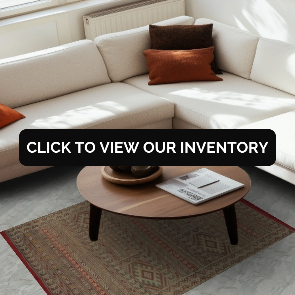 A cream corner sofa with warm orange and brown pillows is bathed in natural sunlight. Beneath the round wooden coffee table lies a small rug with muted geometric motifs in red, tan, and beige. A black “CLICK TO VIEW OUR INVENTORY” button covers the center of the image.