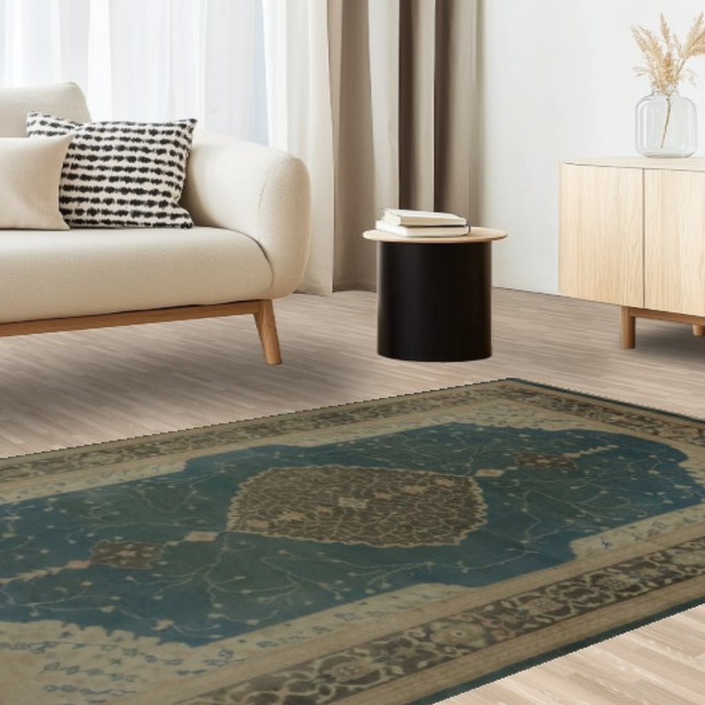 A cozy modern living room features a cream sofa with a patterned pillow, paired with a black accent table and light wood cabinetry. The floor is anchored by a vintage-style blue rug with tan and cream detailing. Soft natural light from sheer curtains brightens the space.