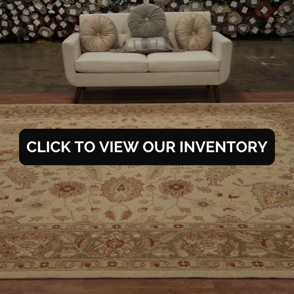 A cream loveseat with decorative round pillows is displayed in a showroom stacked with rolled rugs in the background. A large beige rug with warm floral patterns covers the floor in front of the sofa. A black “CLICK TO VIEW OUR INVENTORY” button stretches across the lower portion.