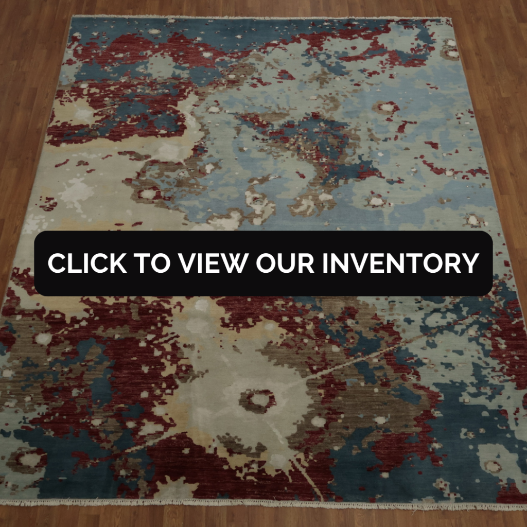 A large abstract rug features swirling patches of blue, cream, red, and taupe, creating a painterly, textured design. The rug sits on a hardwood floor, filling the frame. A black “CLICK TO VIEW OUR INVENTORY” button overlays the center.