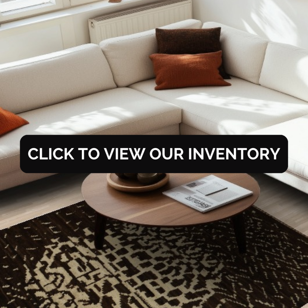 A cream sectional sofa with warm orange pillows sits in bright natural light. Beneath a round wooden coffee table lies a dark brown and beige rug with an irregular geometric pattern. A black overlay reads “CLICK TO VIEW OUR INVENTORY.”