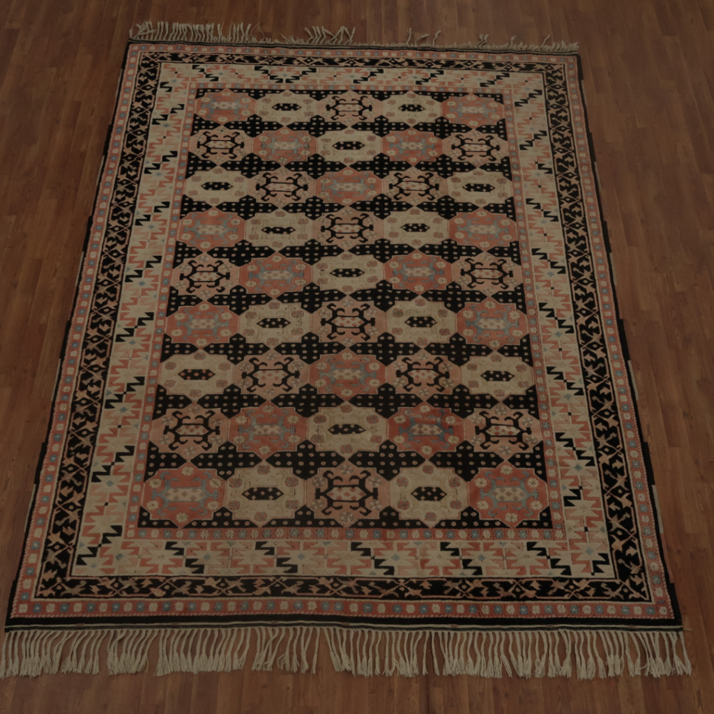 A large flatwoven rug displays a repeating geometric pattern in peach, turquoise, cream, and black. Decorative borders frame the central design, and fringe tassels line the top and bottom edges. The rug sits on a warm wooden floor, photographed in full view.