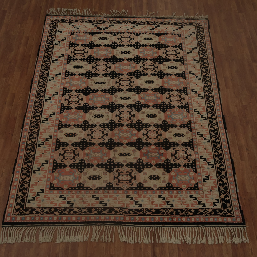 A large flatwoven rug displays a repeating geometric pattern in peach, turquoise, cream, and black. Decorative borders frame the central design, and fringe tassels line the top and bottom edges. The rug sits on a warm wooden floor, photographed in full view.