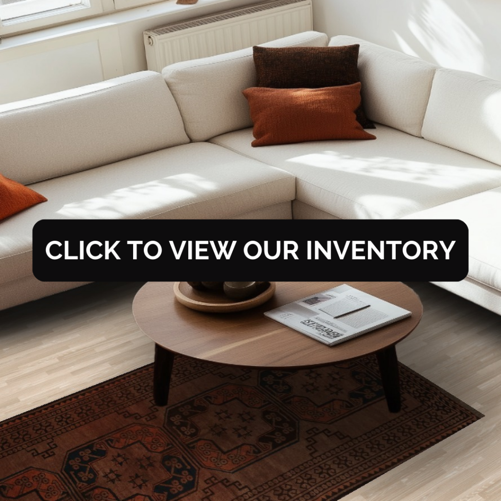 A bright living room features a cream-colored sectional sofa with rust and brown accent pillows, paired with a round wooden coffee table. Beneath the table is a patterned rust-colored rug with geometric motifs. A black button with white text reading “CLICK TO VIEW OUR INVENTORY” is centered over the scene.
