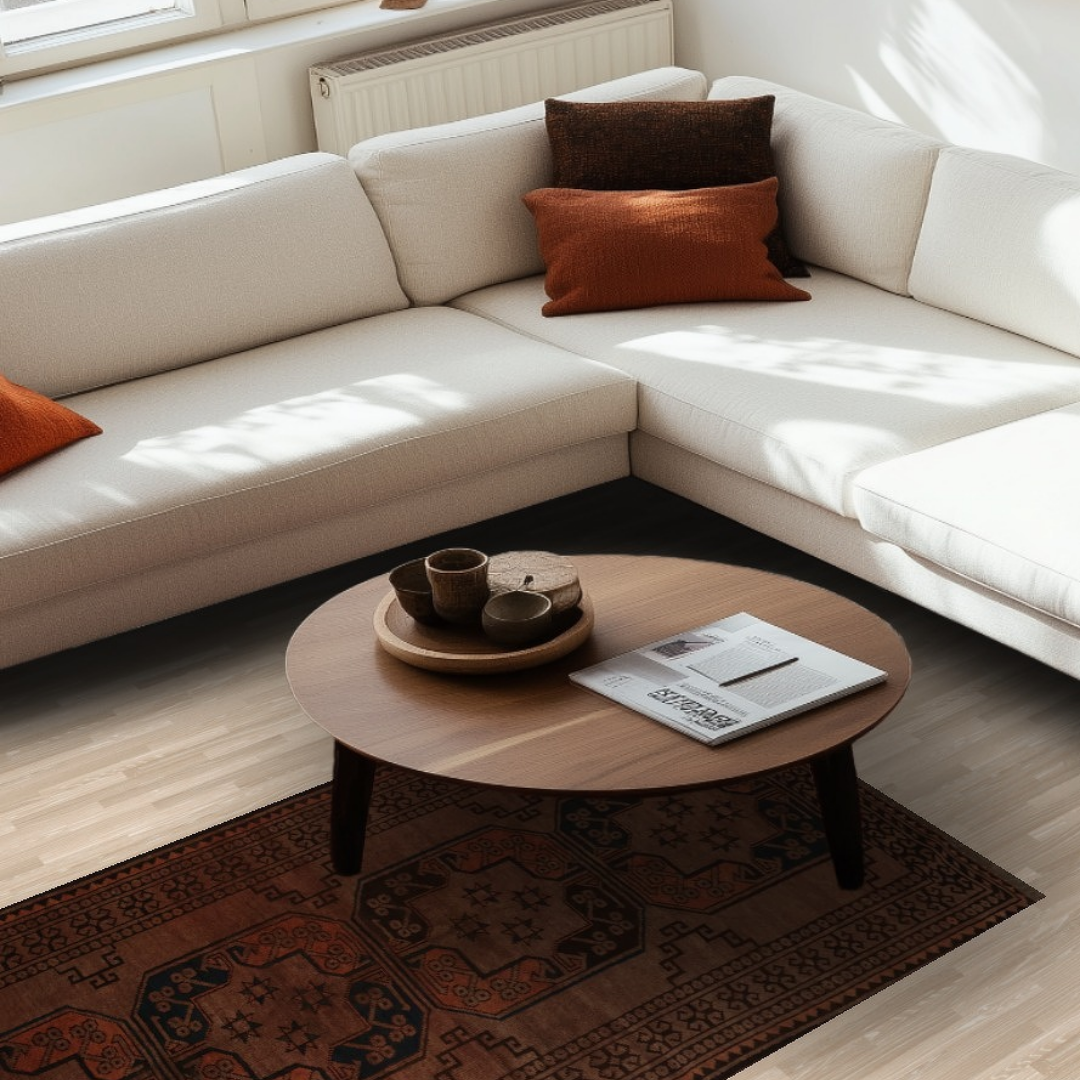 A modern living room shows a cream-colored sectional sofa accented with rust and brown pillows. Sunlight filters in from the window, highlighting a round wooden coffee table holding a tray and open magazine. A geometric rust-colored rug anchors the space beneath the table.