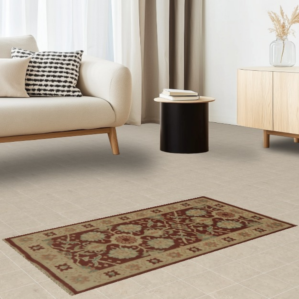 A minimalist living room features a cream sofa with a black-and-white patterned pillow and a small black side table holding a few books. A long, narrow rug with a red and beige floral pattern stretches across the tiled floor. A light wood cabinet with a glass vase of dried stems sits against the wall, completing the clean, modern look.