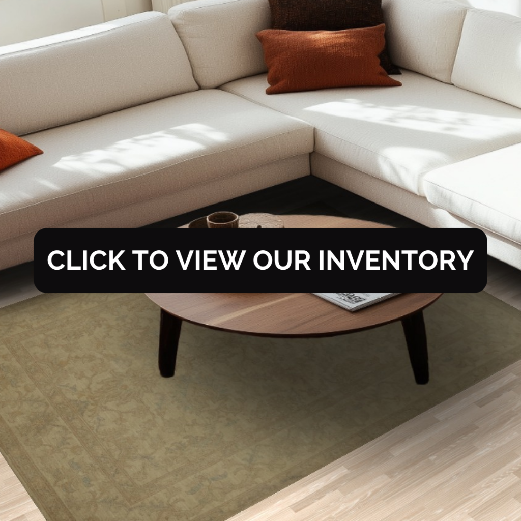A bright living room with a cream-colored sectional sofa and warm-toned accent pillows is shown in soft natural light. A subtle green-beige rug lies beneath a round wooden coffee table holding a tray and magazine. A black button with white “CLICK TO VIEW OUR INVENTORY” text is centered over the rug.