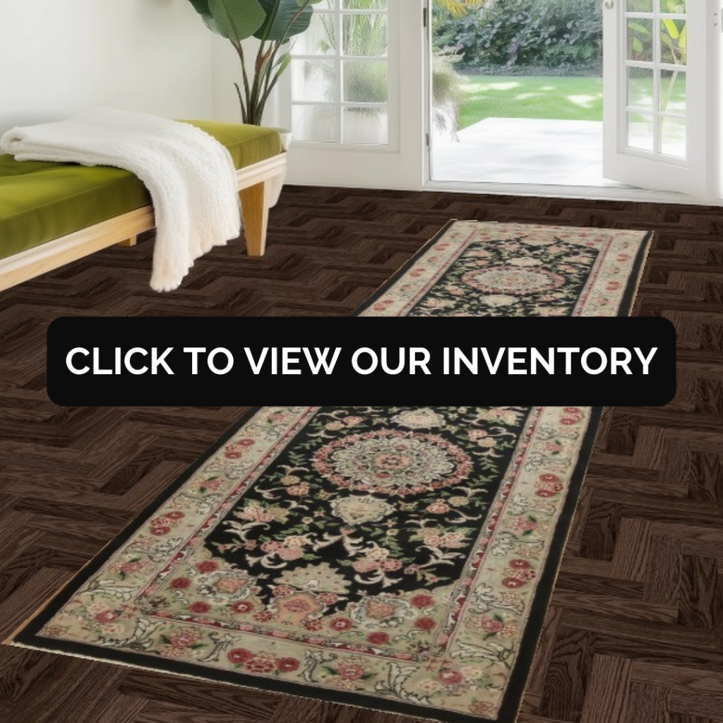 A sunlit entryway features a wooden bench with a green cushion and a tall leafy plant near glass doors. A narrow runner rug with a dark background and detailed floral motifs extends along the floor. The space feels calm, airy, and welcoming.
