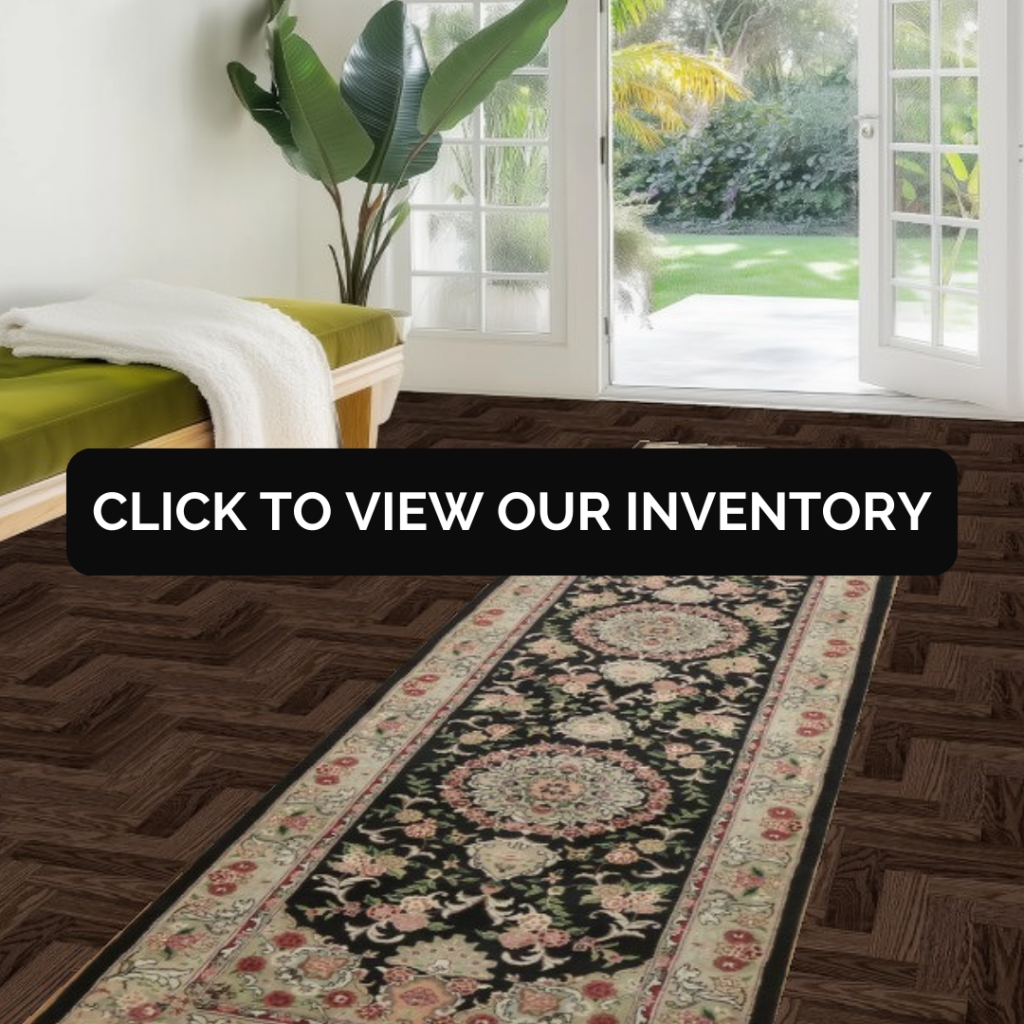 A bright entryway features a wooden bench with a green cushion and a folded white throw near glass doors opening to a garden. A long runner rug with a dark central field and ornate floral medallions stretches across the parquet floor. A black button reading “CLICK TO VIEW OUR INVENTORY” is centered over the image.