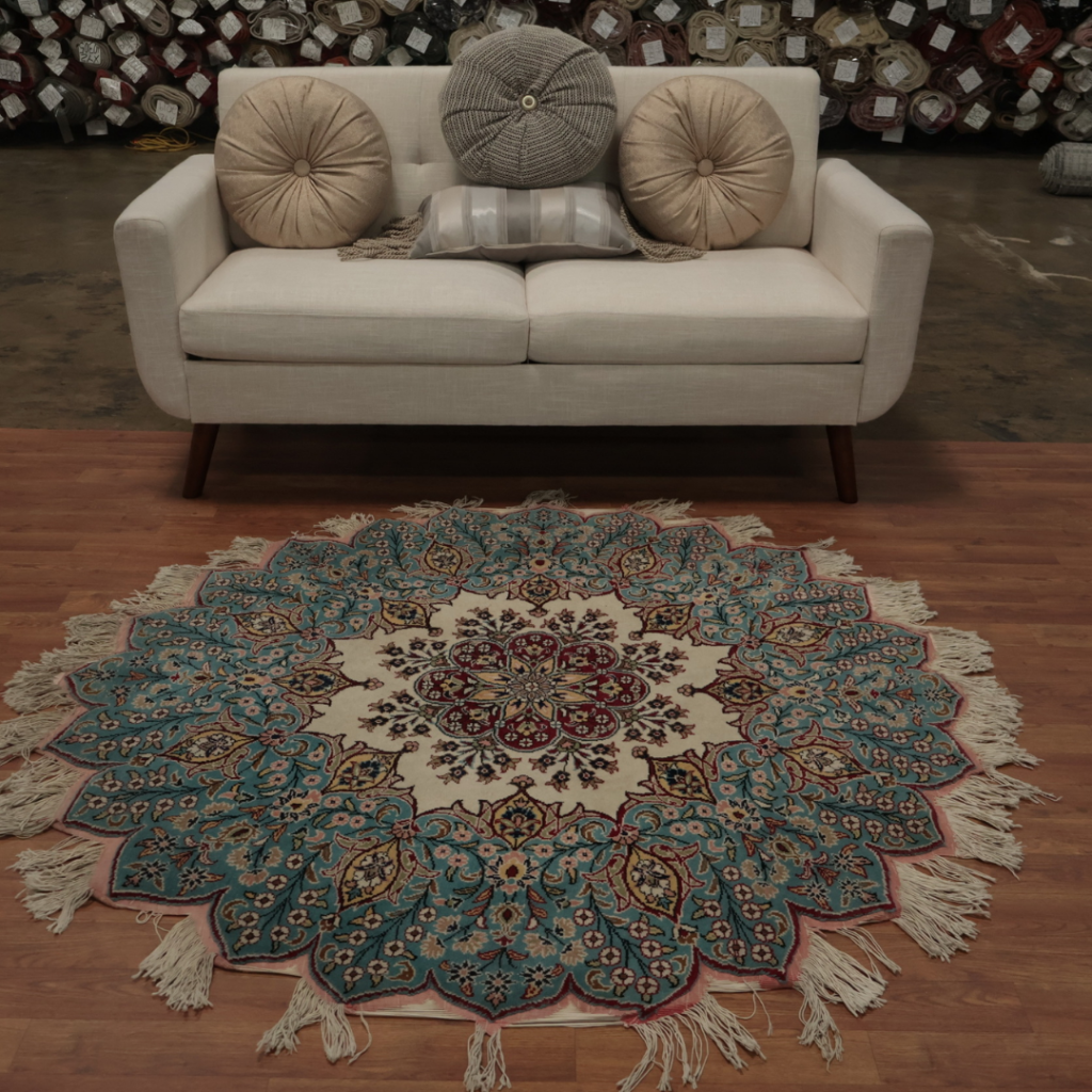 A styled living room scene featuring a light upholstered sofa positioned behind a circular, traditional rug with intricate teal and red detailing. The rug is centered on hardwood flooring and framed by stacked rugs in the background.