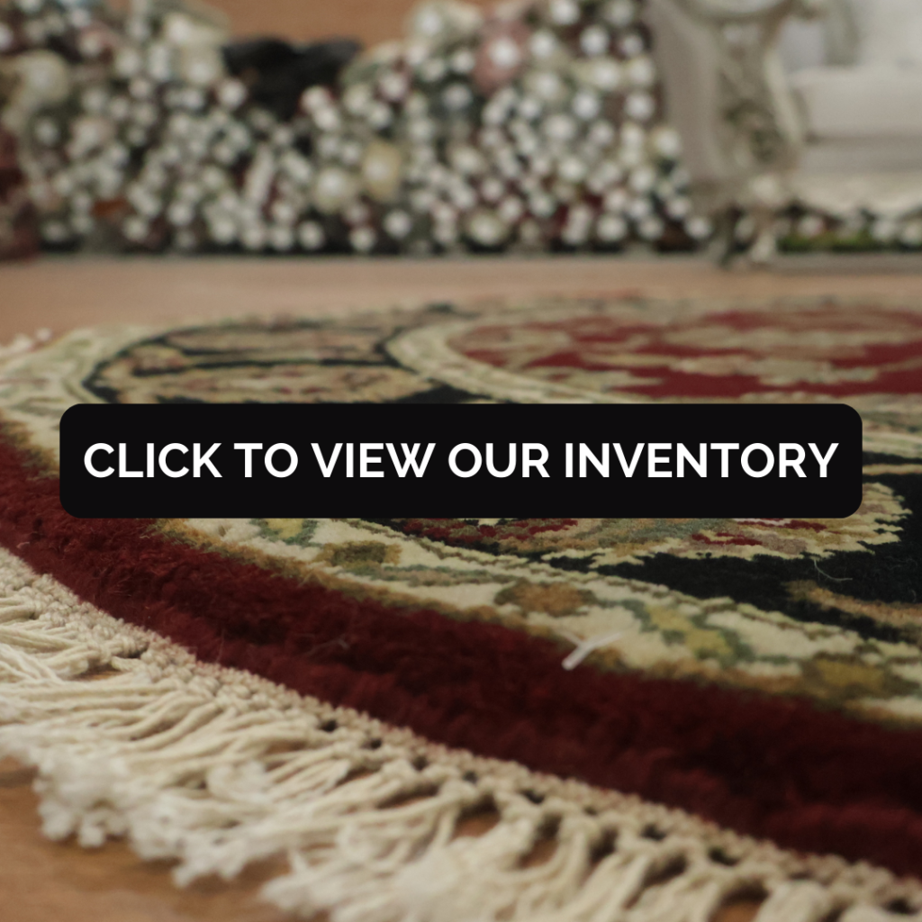 Close-up, low-angle view of a patterned area rug showing plush texture, deep red accents, and cream fringe along the edge. Rolled rugs are softly blurred in the background, suggesting a showroom setting.