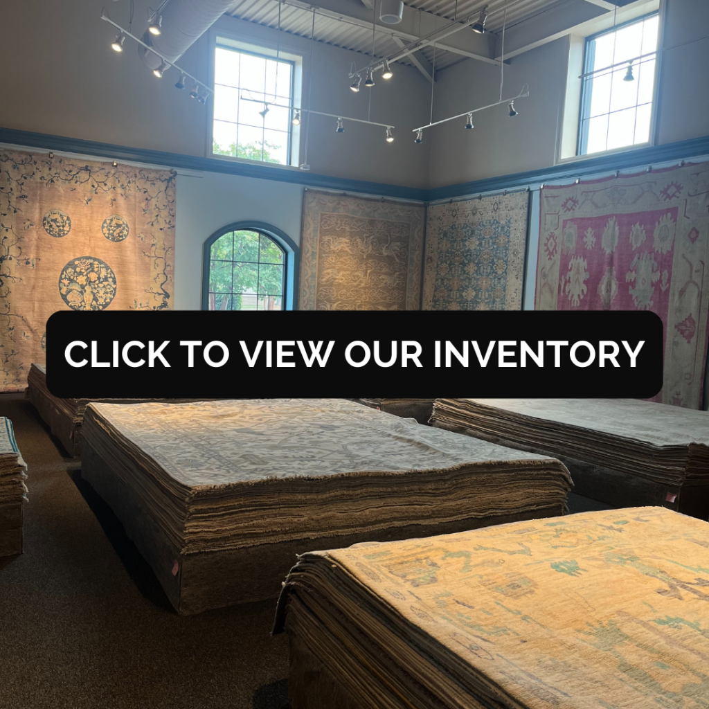 Interior of a rug showroom with large stacks of folded area rugs arranged in rows. Decorative rugs hang on the walls beneath tall windows and ceiling track lighting.