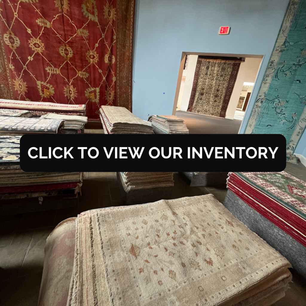 Rug showroom interior showing multiple stacks of folded rugs in neutral and muted tones. Large patterned rugs hang on the walls, creating a gallery-like display.