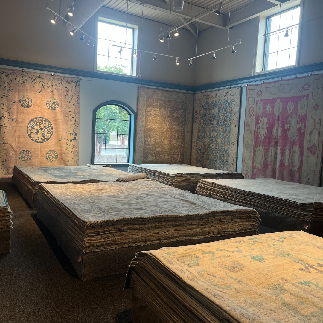A bright showroom space filled with neatly stacked rugs laid flat on low platforms. Tall windows, overhead lighting, and hanging rugs highlight the variety of patterns and textures.