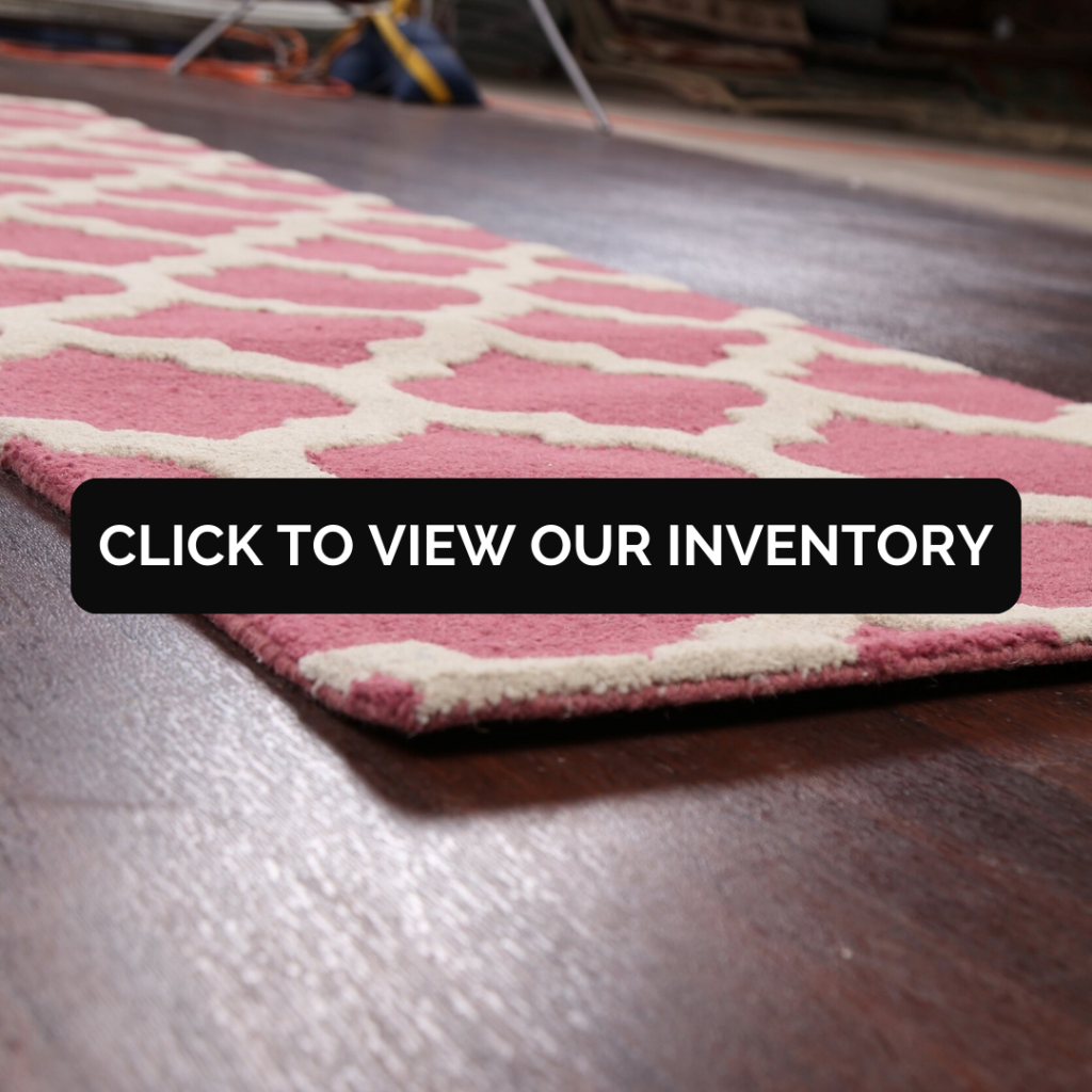 Low-angle close-up of a pink and cream patterned area rug showing its plush pile and stitched edge. The rug rests on a dark wood floor with a softly blurred showroom background.