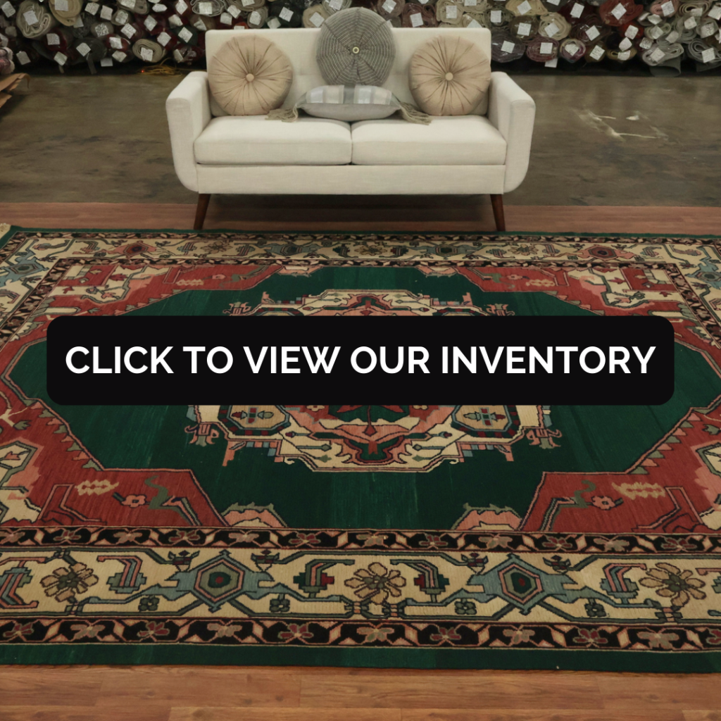 A traditional area rug with a dark green center and ornate red medallion fills the foreground of a showroom setting. Intricate borders in cream, teal, and black surround the central motif. A black “CLICK TO VIEW OUR INVENTORY” button is centered over the rug.
