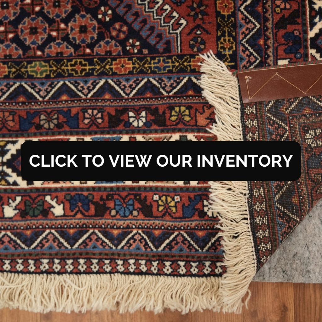 A close-up view shows the corner of a richly colored rug featuring dense geometric and floral motifs in navy, rust, gold, and cream. White fringe lines the edge, and a leather tag rests near the corner. A black button reading “CLICK TO VIEW OUR INVENTORY” overlays the center of the image.