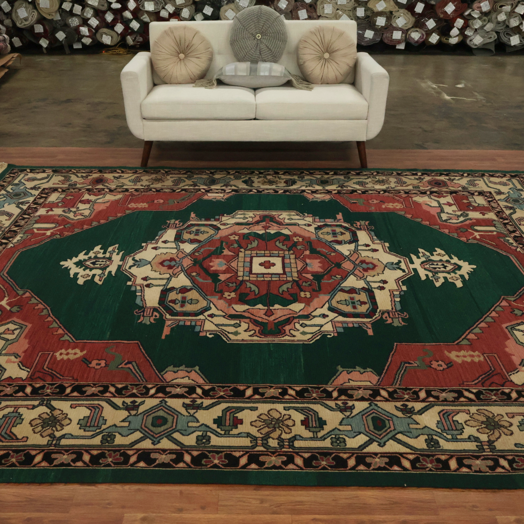 A large traditional rug with a deep green field and red central medallion is displayed on a hardwood floor in a rug showroom. Cream, teal, and red geometric borders frame the design. A white loveseat with round accent pillows sits behind the rug, with rolled rugs stacked in the background.