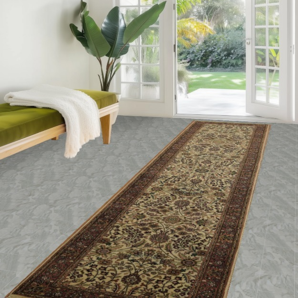 A light-filled entryway shows a wooden bench with a green cushion and large leafy plant near glass doors leading outside. A narrow traditional runner rug with intricate floral motifs in beige, brown, and red extends along the floor. The space feels airy, calm, and welcoming.
