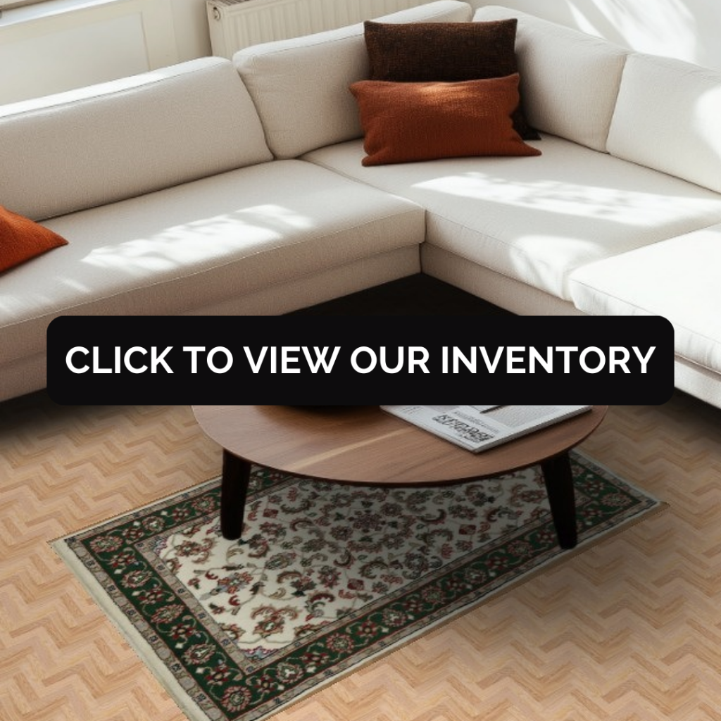 A cream-colored sectional sofa with rust and brown accent pillows sits in a sunlit living room. Beneath a round wooden coffee table is a small traditional rug with a cream field and green and red floral border. A black button reading “CLICK TO VIEW OUR INVENTORY” overlays the center of the image.