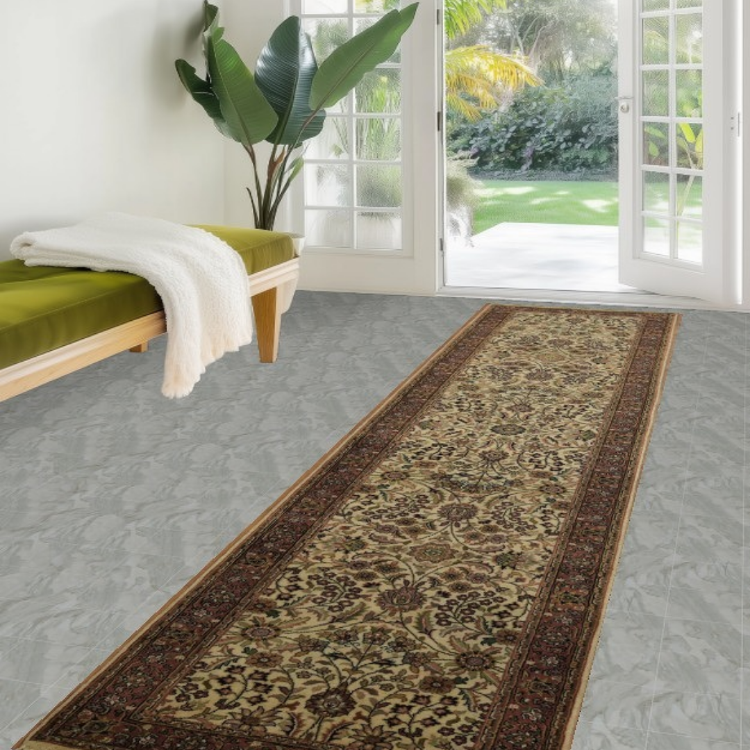 A light-filled entryway shows a wooden bench with a green cushion and large leafy plant near glass doors leading outside. A narrow traditional runner rug with intricate floral motifs in beige, brown, and red extends along the floor. The space feels airy, calm, and welcoming.