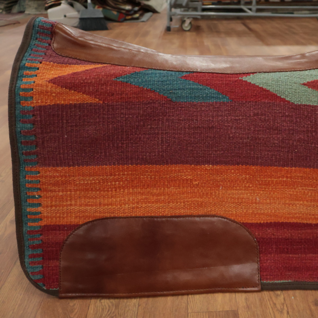 A close-up shows a woven saddle blanket featuring horizontal bands of rust red, burnt orange, teal, and sage green. Smooth brown leather reinforcements are stitched along the top edge and lower corner. The blanket rests on a hardwood floor in a workshop or showroom setting.