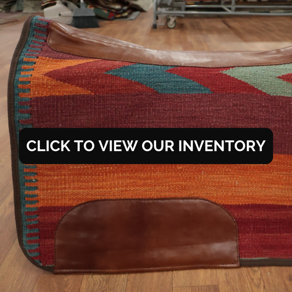 A textured saddle blanket with warm red and orange stripes and teal geometric accents is shown in close detail. Brown leather panels reinforce the edges and corners. A black button reading “CLICK TO VIEW OUR INVENTORY” overlays the center of the image.