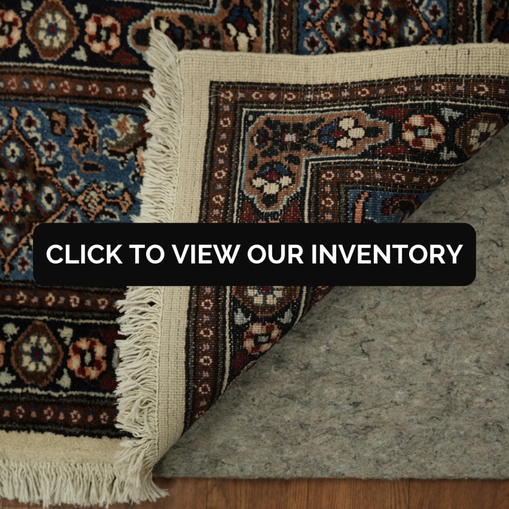 “Close-up of a traditional patterned rug with fringe, partially folded to show backing on a felt rug pad. Black button reads ‘Click to View Our Inventory.’”