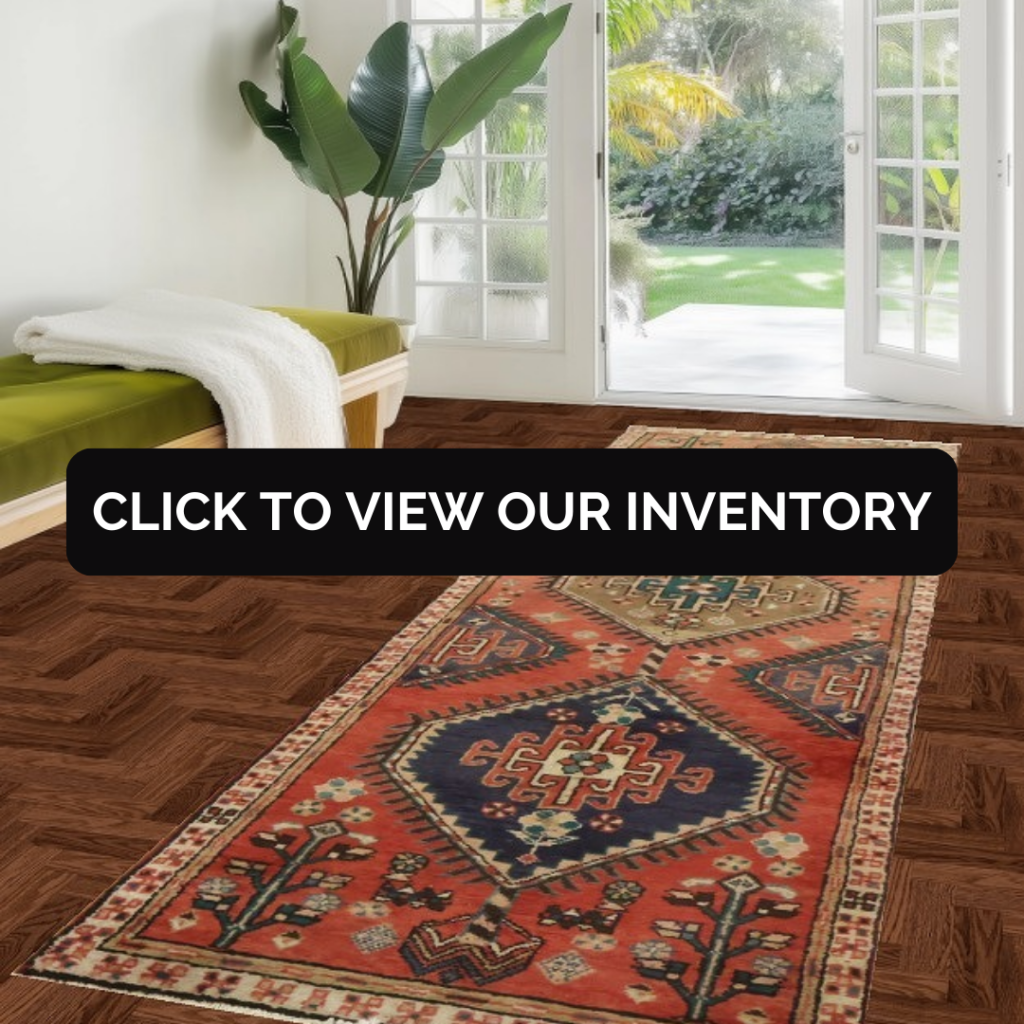 A colorful runner rug with red, navy, and cream geometric patterns lies on a dark herringbone wood floor in a bright room with glass doors opening to a garden. A bench with a green cushion and a large potted plant sit nearby. A black banner across the center reads, “CLICK TO VIEW OUR INVENTORY.”